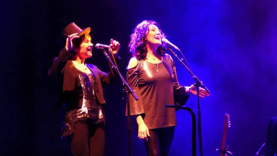 Two women singing on stage with microphones, one wearing a hat and the other with curly hair, illuminated by purple stage lighting.