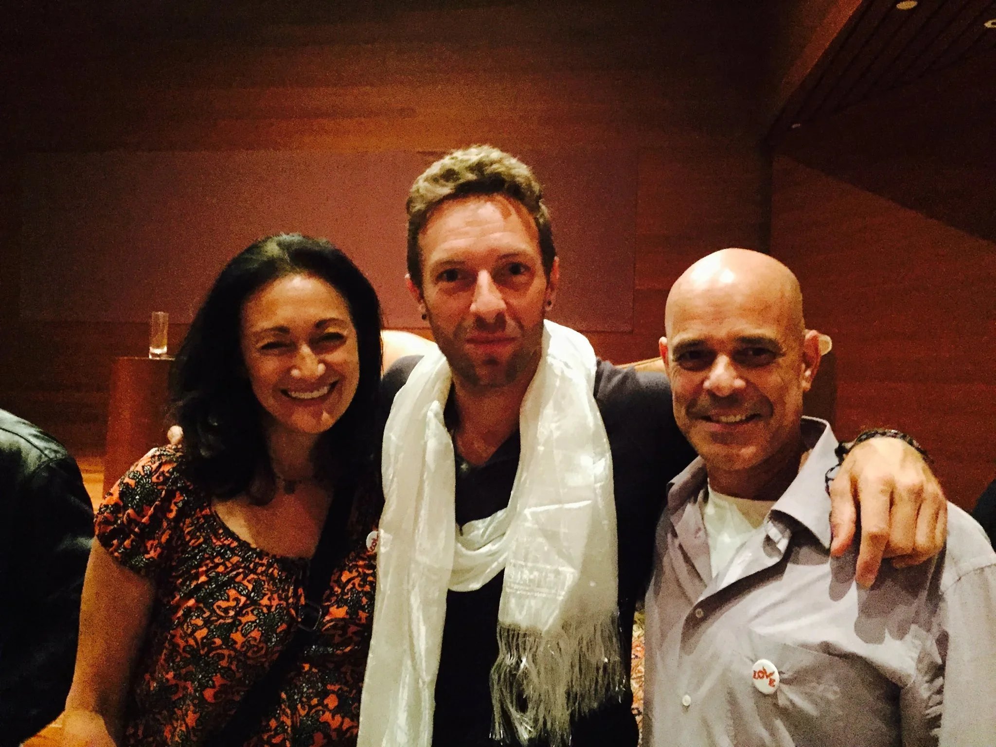 Three people smiling and posing closely together indoors with a dark red wall background. The woman on the left has black hair and is wearing a patterned dress, the man in the middle has short light brown hair and is wearing a white scarf and shirt, 