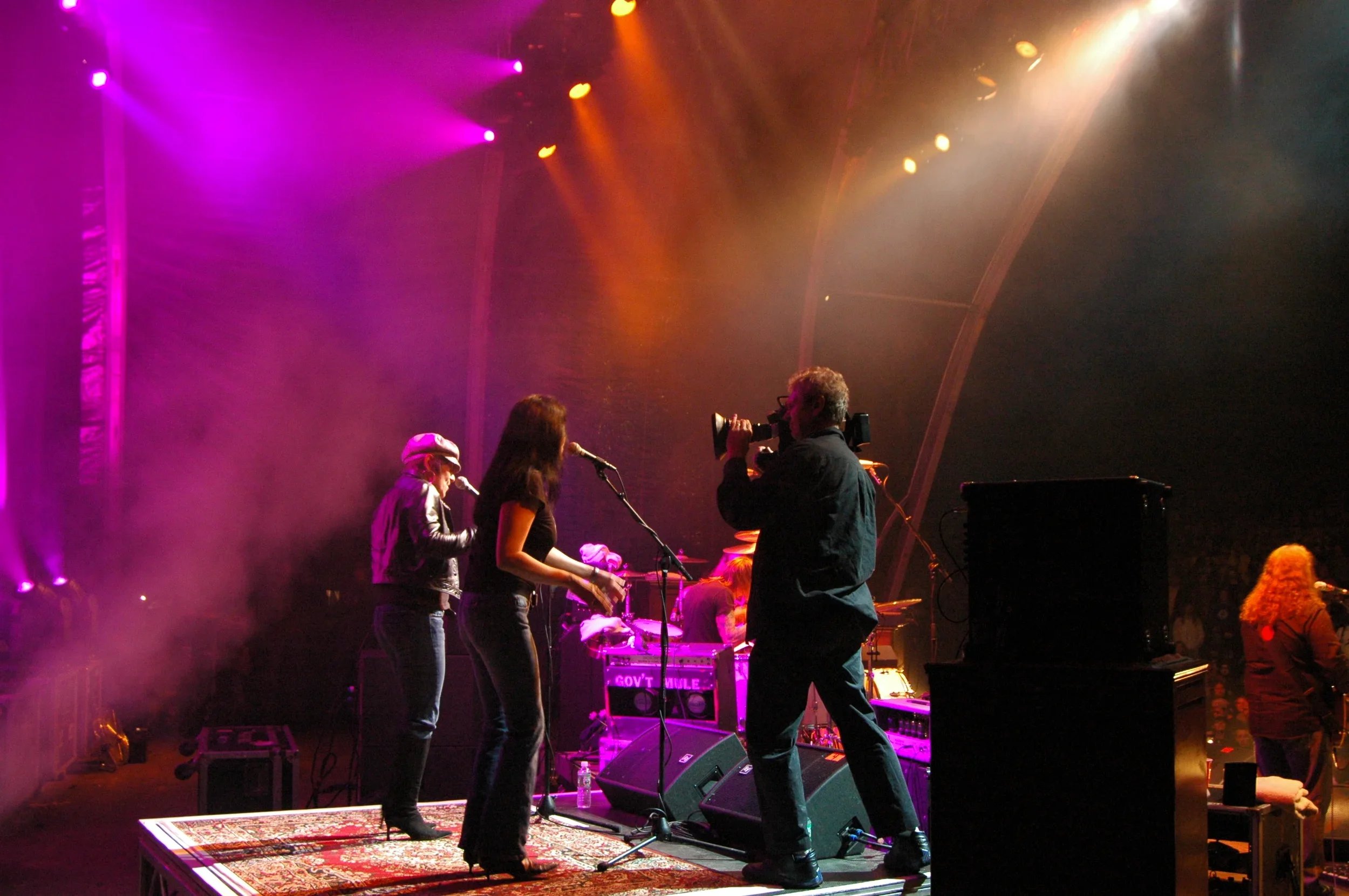 Musicians performing on stage with colorful lighting, including two women singing into microphones and a man filming with a video camera, in a concert venue.