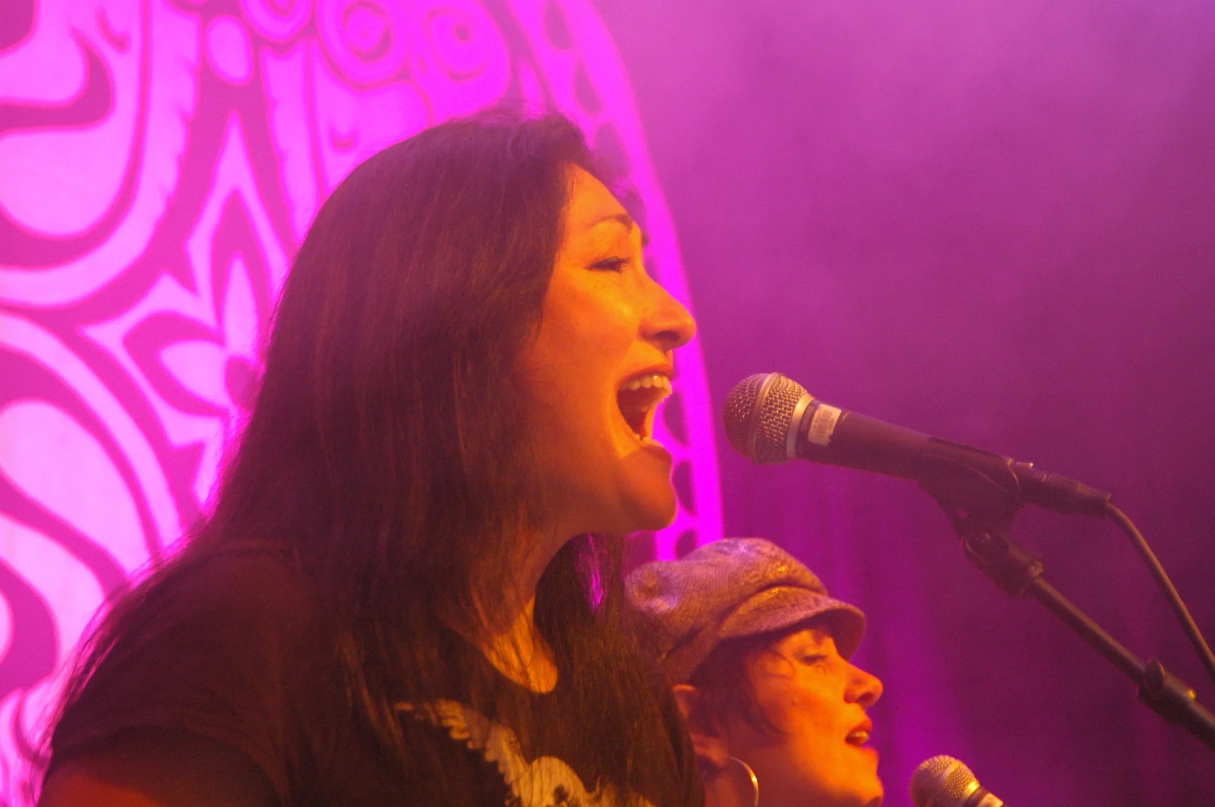 A woman singing into a microphone on stage with a vibrant pink background and artistic wall design.