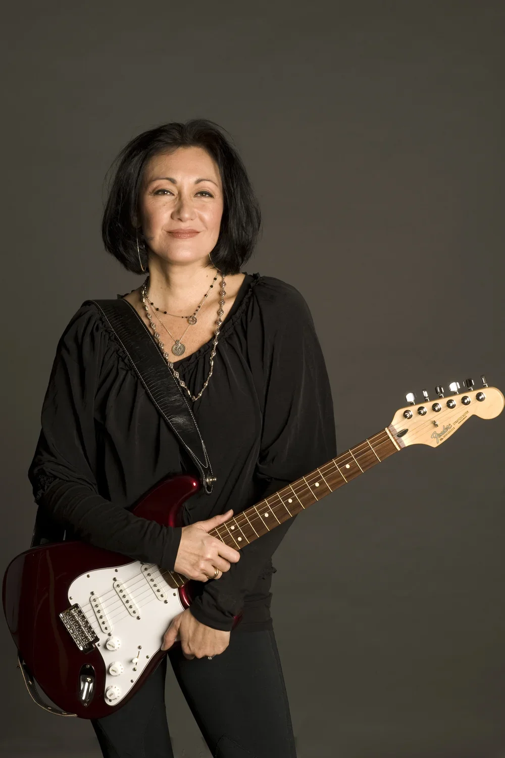 A woman with black hair holding a red and white electric guitar, wearing a black top and jewelry, standing against a gray background.