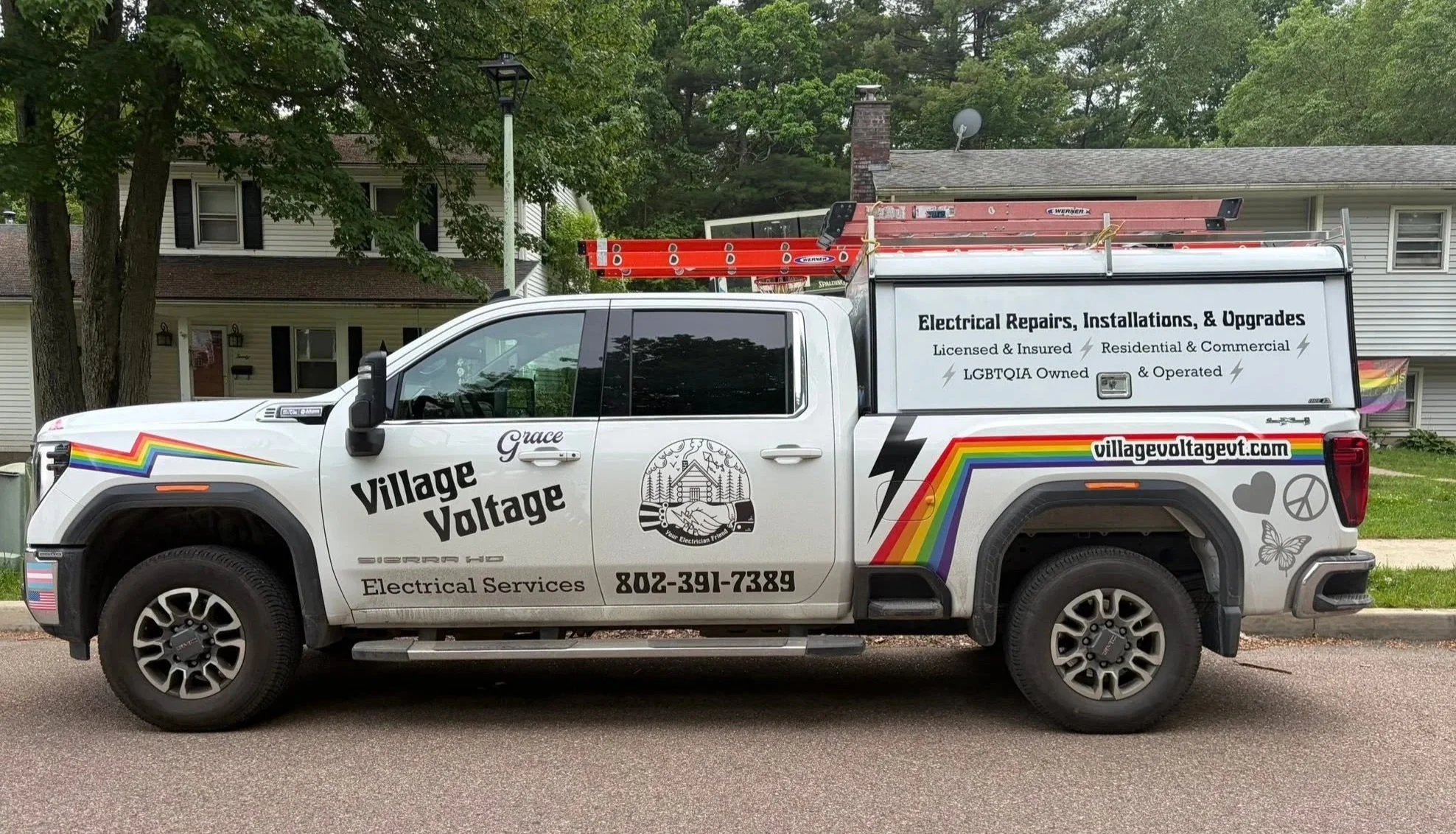 A white service truck with rainbow and peace symbols, advertising electrical services for a company called Village Voltage. The truck has a ladder on top and the company’s logo and contact number on the side. It is parked in front of houses with green trees in the background.