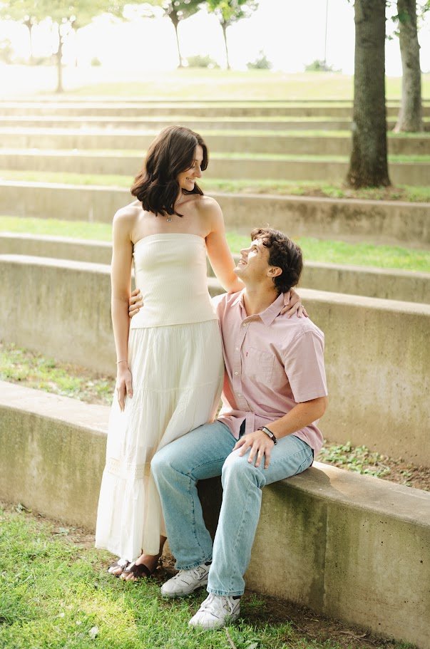 A woman in a white strapless dress and a man in a pink shirt and jeans sitting on outdoor concrete steps, looking at each other happily.