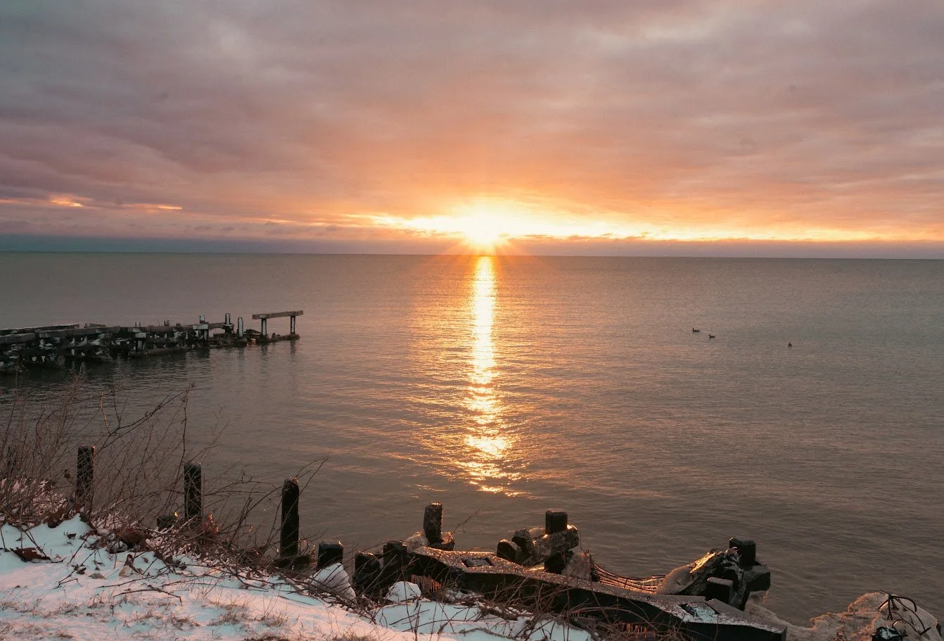 Sunset over a calm ocean with a partially frozen shoreline, wooden posts, and a small dock on the left.