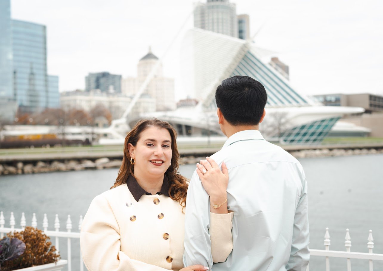 A woman smiling at a man during an outdoor encounter near a river with modern buildings in the background.