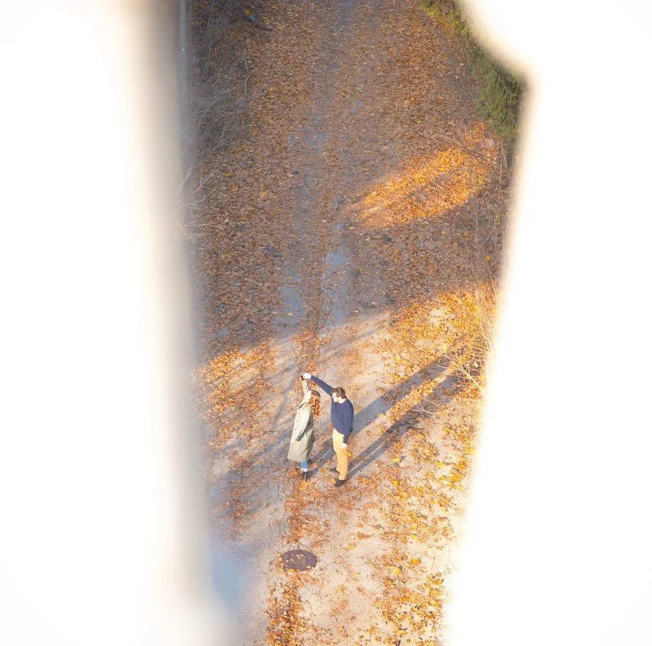 An aerial view of two people standing on a fall sidewalk, holding hands, surrounded by orange autumn leaves.