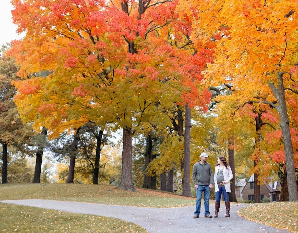 A couple walking on a paved path through a park with trees showcasing vibrant fall foliage in shades of orange, yellow, and red.