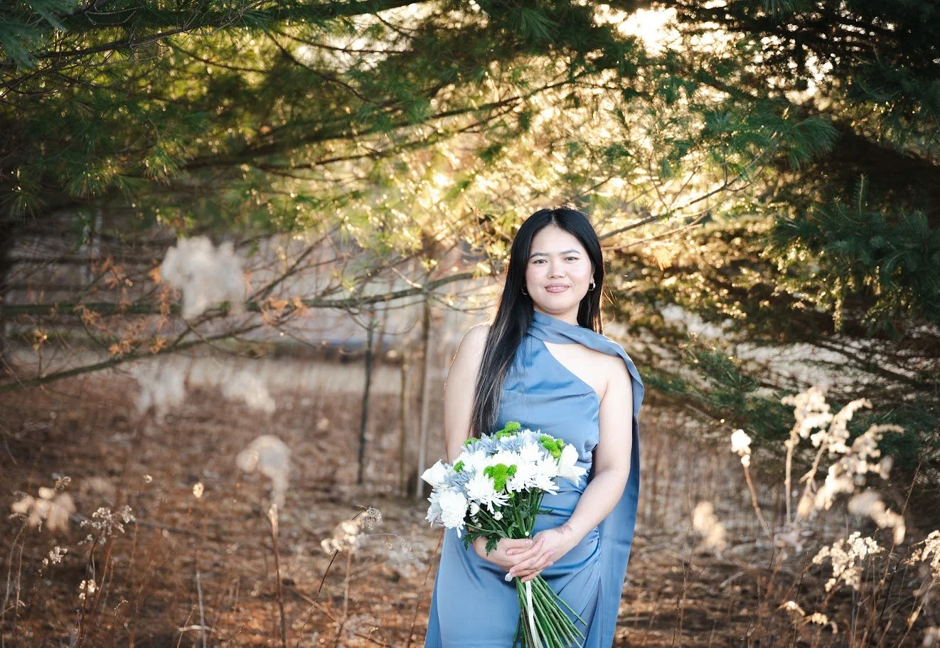A woman in a blue dress holding a bouquet of white and green flowers stands outdoors under a tree with green leaves. The sunlight filters through the branches, creating a warm glow around her.