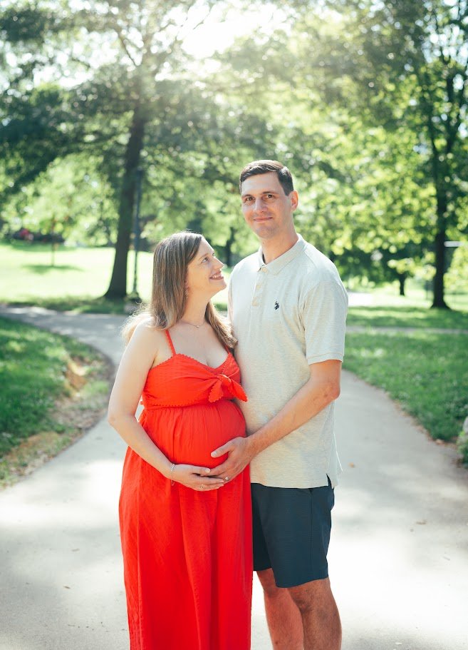 A pregnant woman in a red dress standing next to a man in a beige polo shirt and dark shorts, outdoors on a sunny day with green trees in the background.
