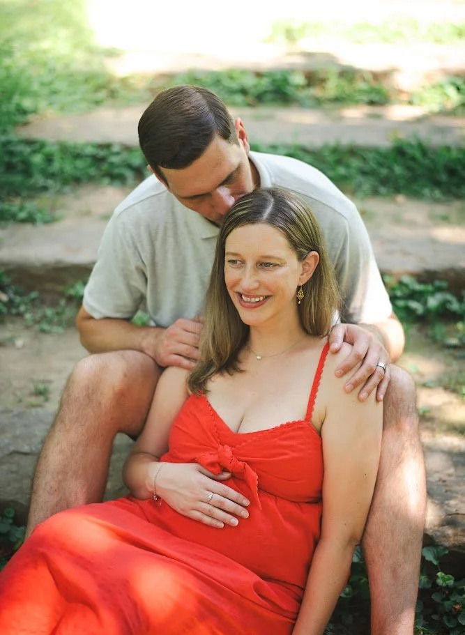 A couple sits outdoors on steps, a woman in a red dress smiling, a man leaning in with his hands on her shoulders and stomach, surrounded by greenery.
