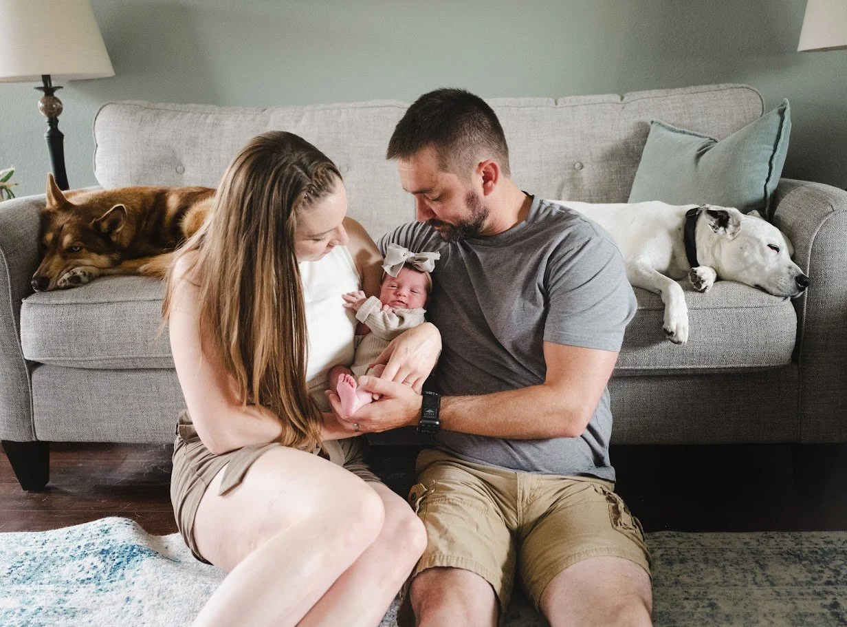A family with a newborn baby sitting on the floor, with two dogs lying on the couch behind them.