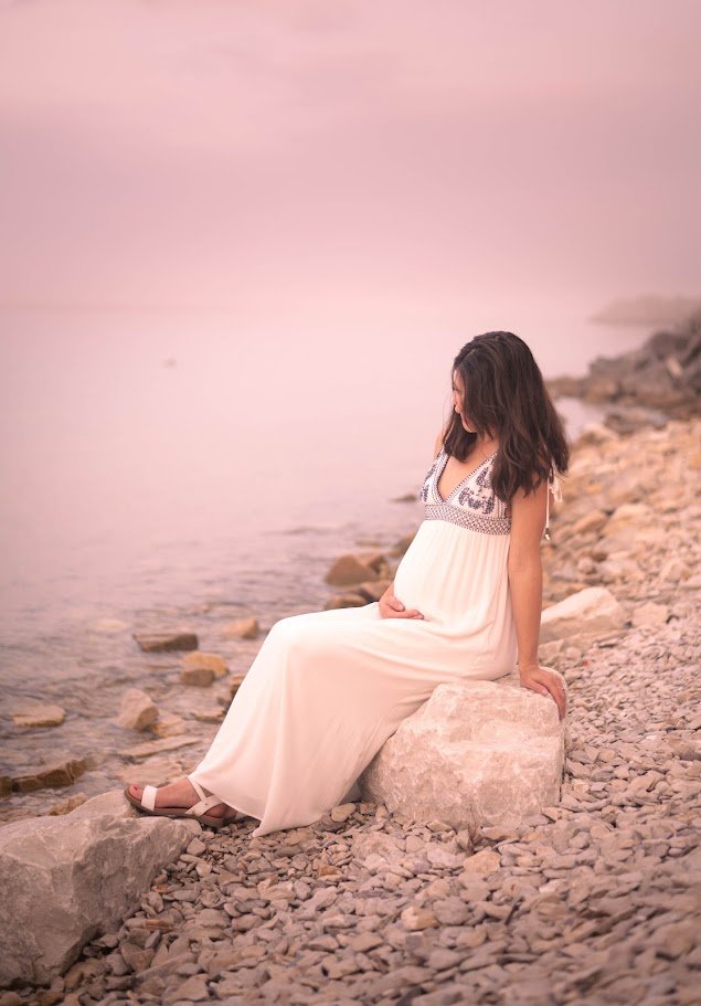 A woman in a white dress sitting on a rock by a rocky lakeside or shoreline, with a calm water and pinkish sky in the background.