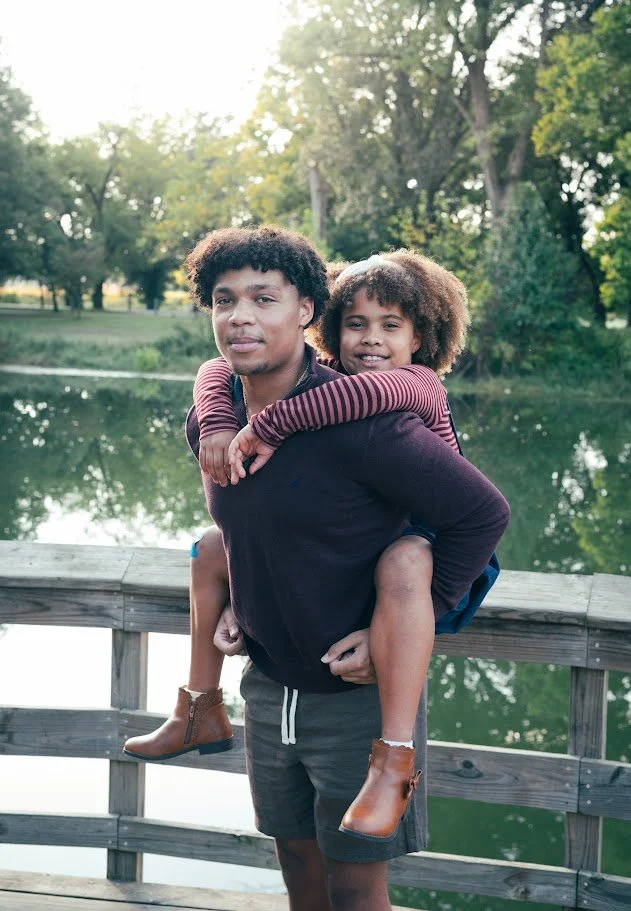 A young man giving a piggyback ride to a girl with curly hair, both smiling, on a wooden dock by a lake surrounded by trees.