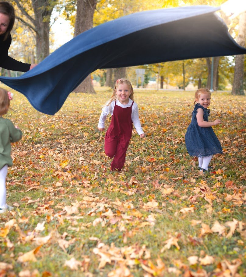 Children playing outdoors in fall leaves while a person lifts a large blue blanket or cloth above them.