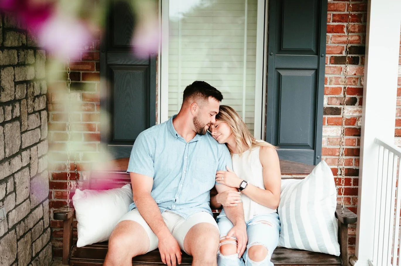 A couple sitting on a porch swing, smiling and leaning their foreheads together, with brick walls and windows in the background.