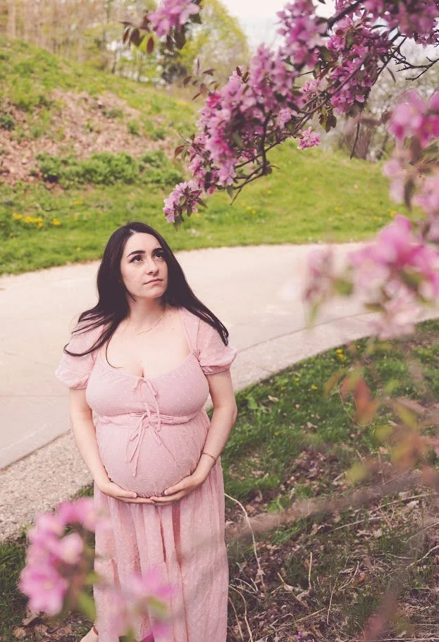 A pregnant woman with long dark hair standing outdoors on a curved path, gently holding her baby bump, surrounded by blooming pink cherry blossom trees and green grass.