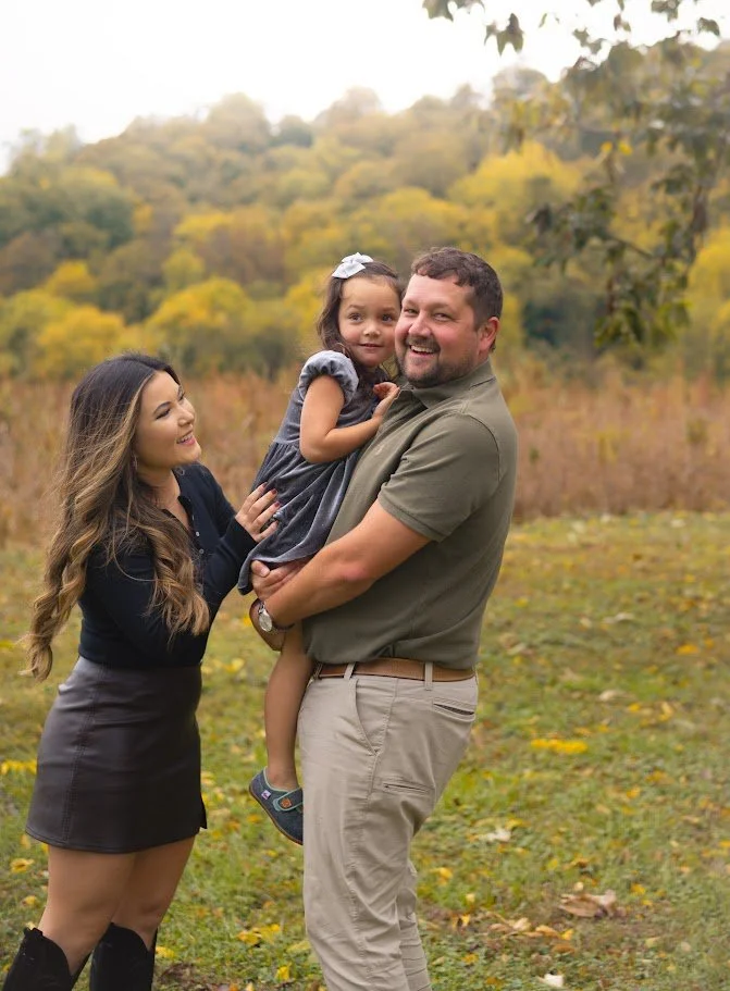 A family of three standing outdoors in a park with autumn foliage, smiling and looking at each other. The man is holding a young girl, and a woman is touching the girl's arm.