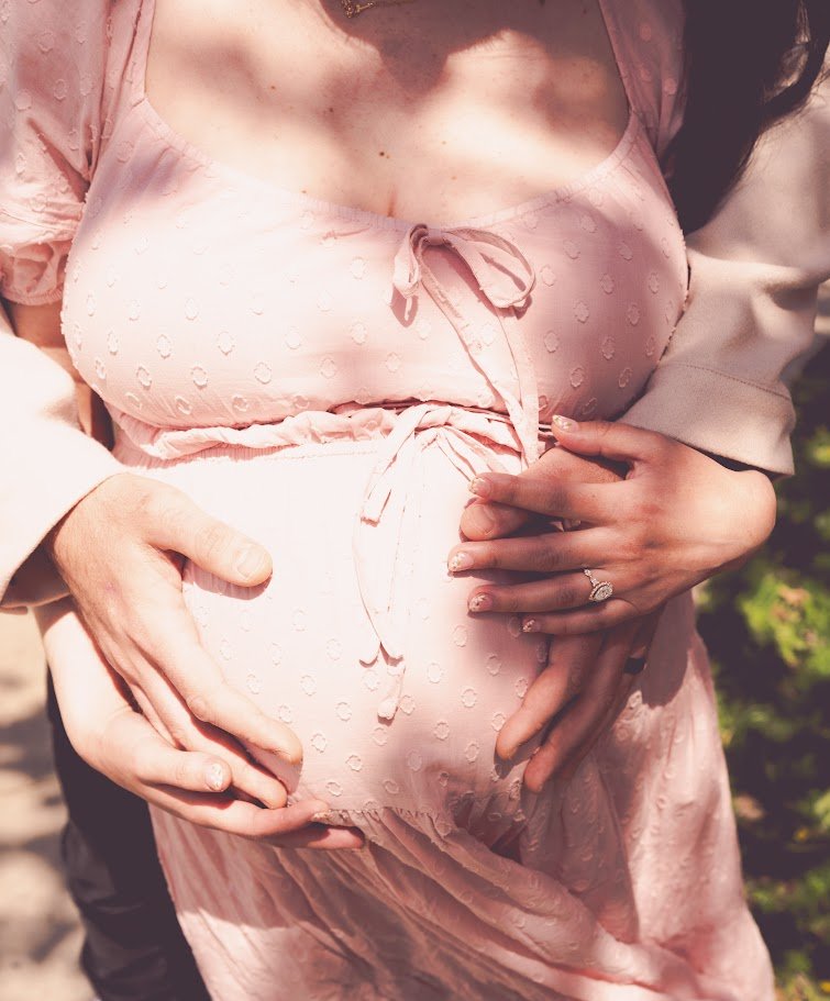 A pregnant woman in a pink dress being gently held by a partner's hands, with an engagement ring visible, outdoors.
