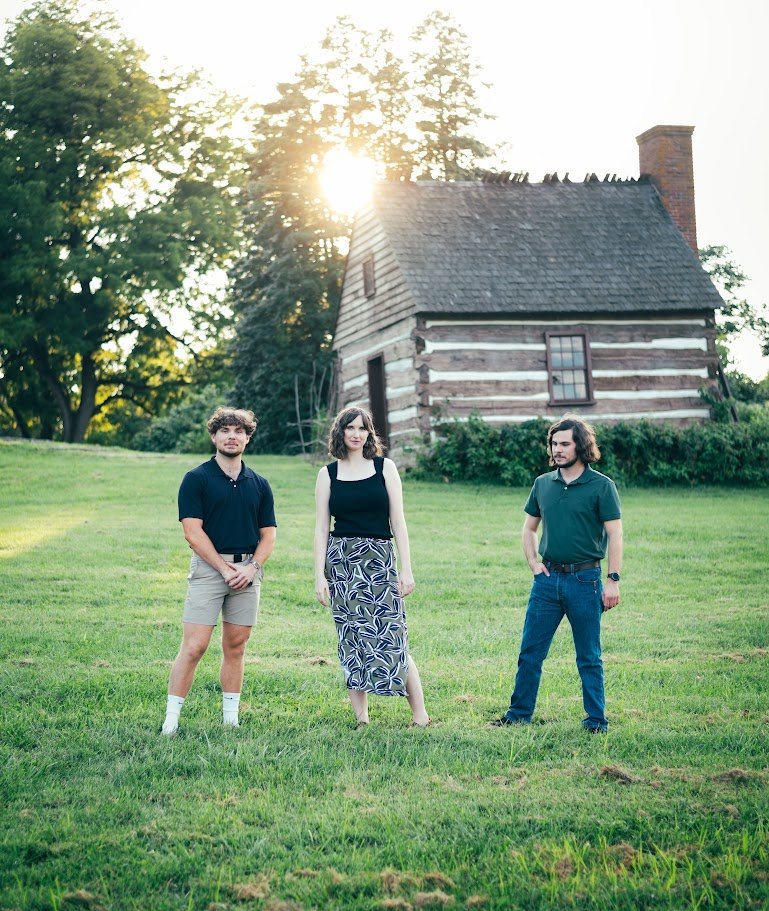 Three young adults standing on a grassy yard in front of a rustic wooden house, with trees and sunlight in the background.