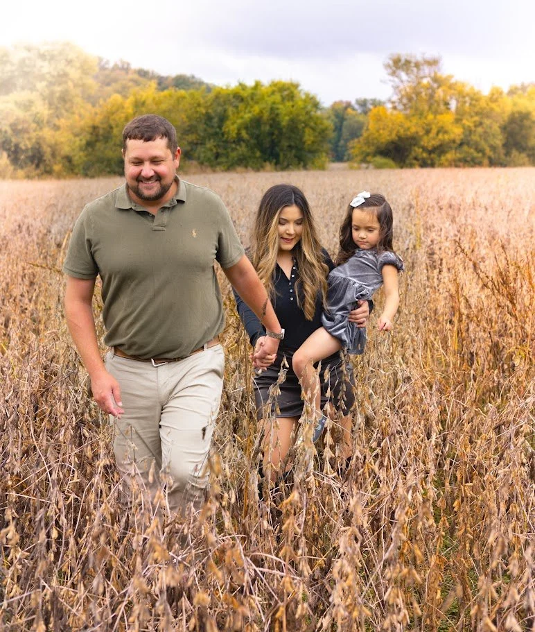 Family walking through a dry, grassy field with trees in the background, enjoying outdoors during autumn.