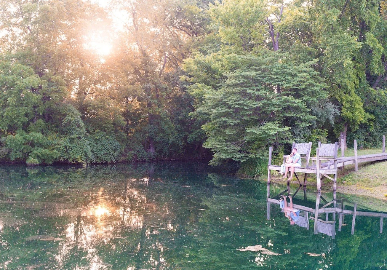 A man sitting on a wooden dock by a calm river, surrounded by lush green trees, with the reflection of the man and trees visible on the water, and sunlight filtering through the leaves.