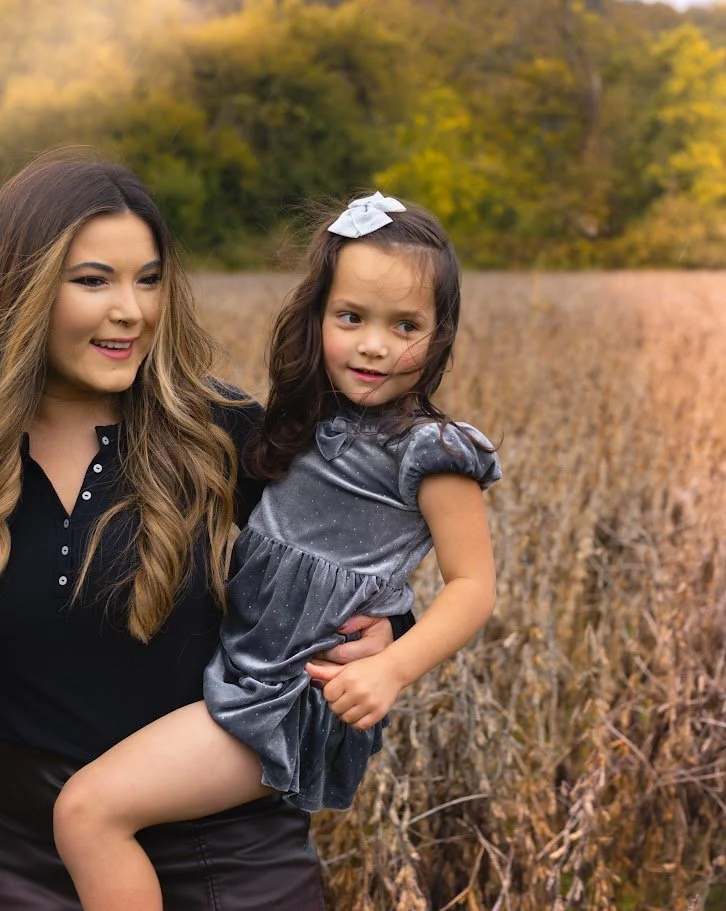 A woman holding a young girl in a field with yellow and orange trees in the background during fall.