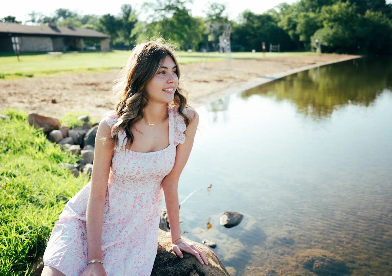 Young woman in a pink floral dress sitting on a rock by a river, smiling and looking to the side on a sunny day.