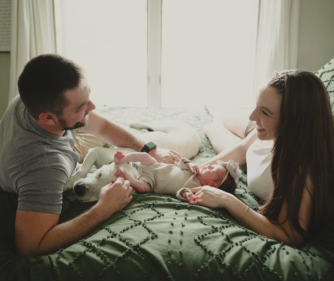 A couple with a newborn baby and a dog on a bed in a bright bedroom, smiling and looking at each other.