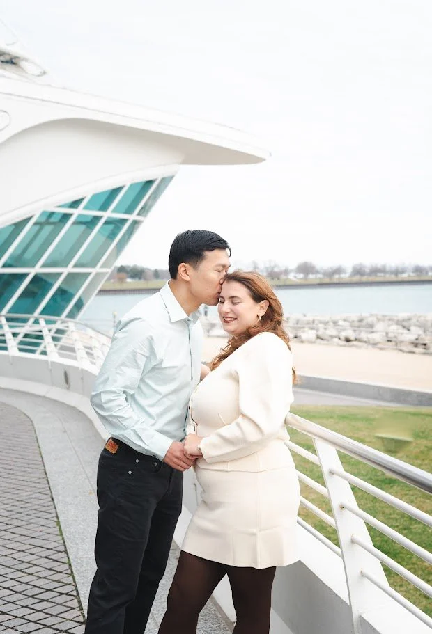 A pregnant woman with red hair wearing a white dress and black tights is smiling while a man with black hair wearing a white shirt and black pants kisses her on the forehead. They are outdoors near a waterfront and modern architectural building with 