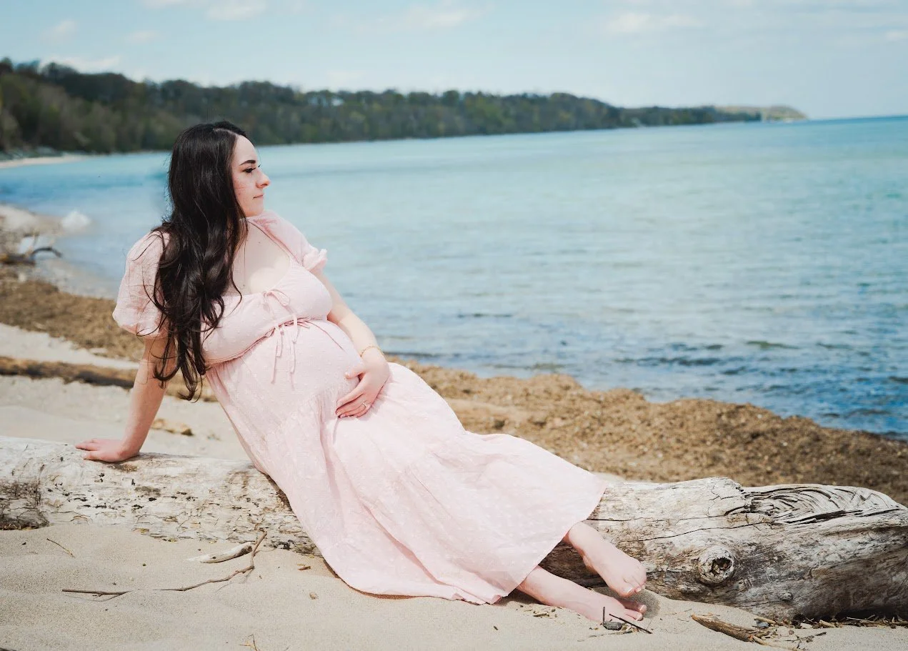 Pregnant woman in a pink dress sitting on driftwood on a beach near the water, with trees and sky in the background.