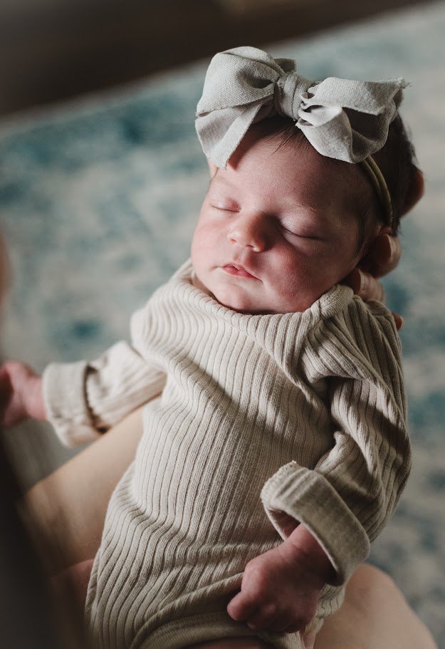 A baby with a large gray bow on her head, sleeping peacefully in a cream-colored, long-sleeved ribbed outfit.