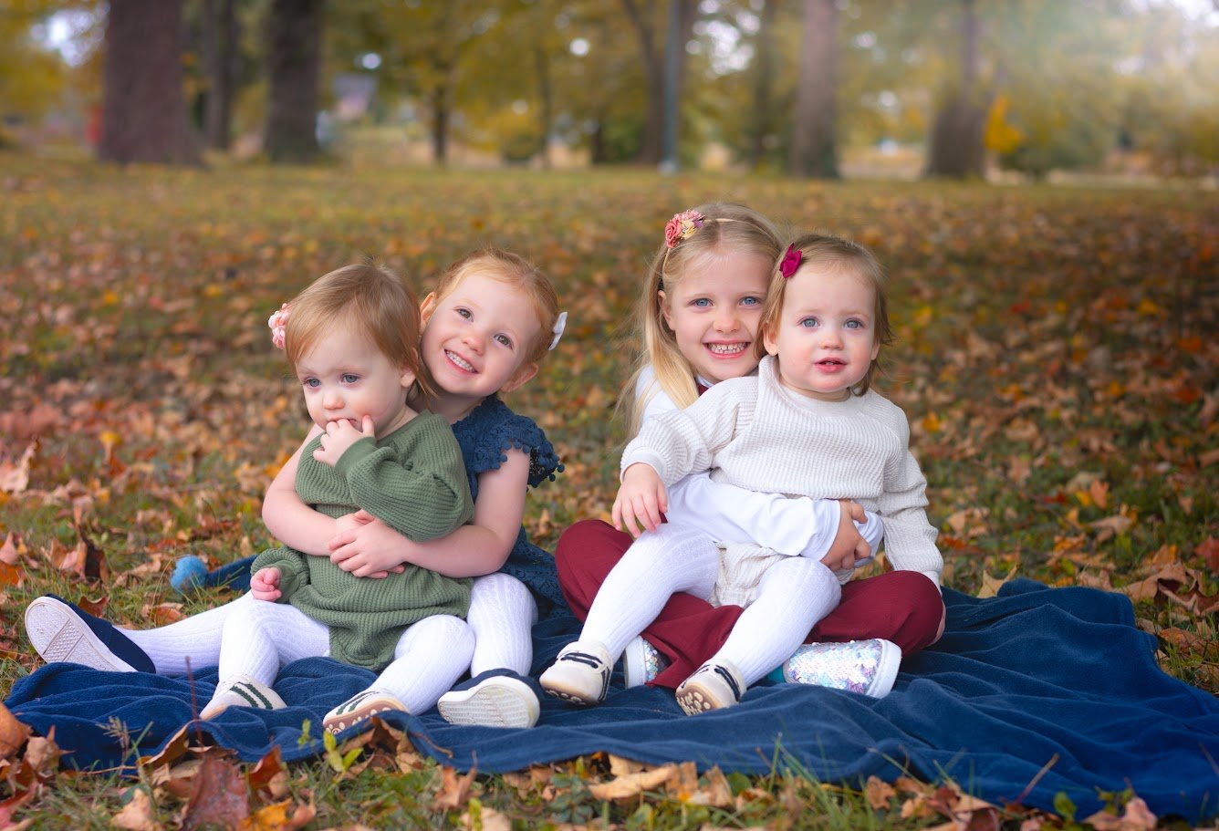 Four young girls sitting on a blanket in a park during autumn, surrounded by fallen leaves and trees.