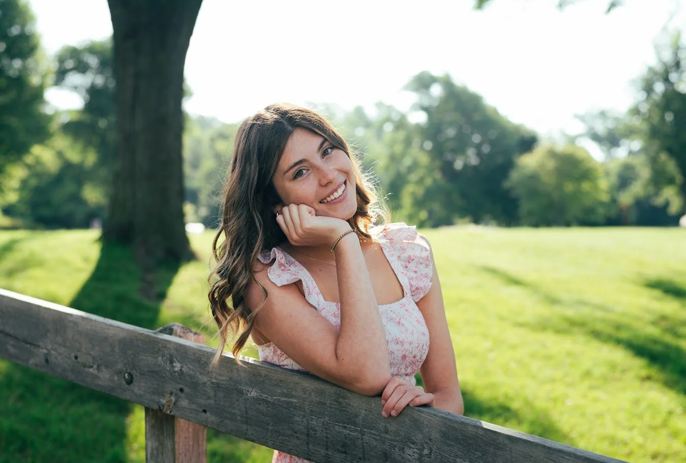 A young woman with wavy brown hair wearing a pink floral dress, leaning on a wooden fence in a park, smiling at the camera with greenery and trees in the background.