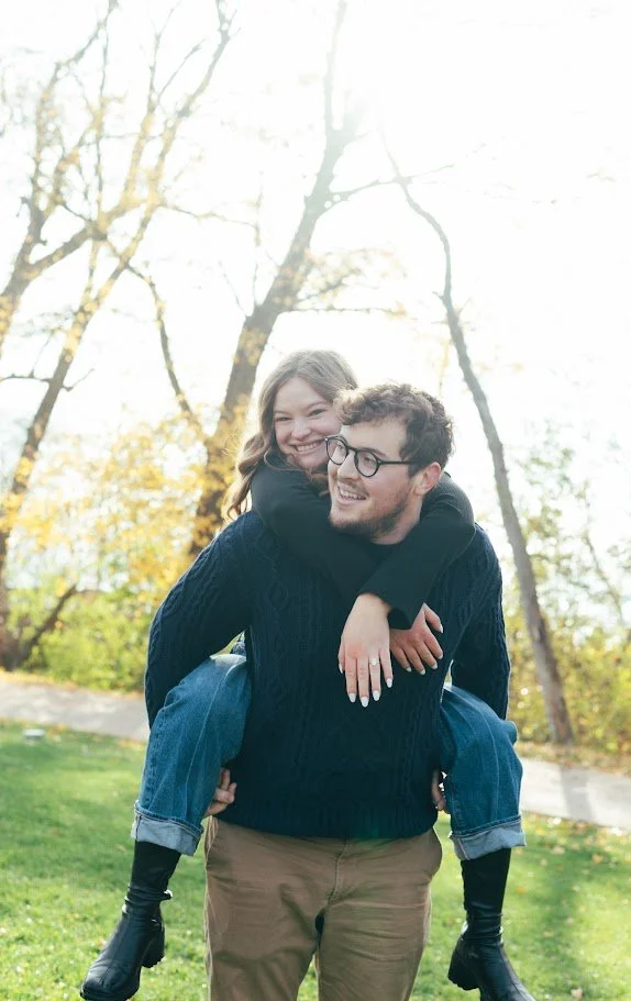 A smiling woman and man enjoying a piggyback ride outdoors in autumn, with trees and fallen leaves in the background.