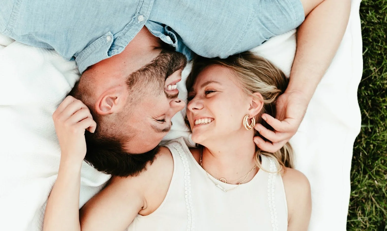 A couple laying on a white blanket outdoors, smiling and touching foreheads, with green grass visible on the side.