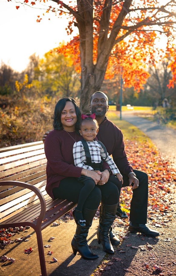 Family of three sitting on a park bench during autumn, with colorful fall leaves and trees in the background, smiling and enjoying the day.