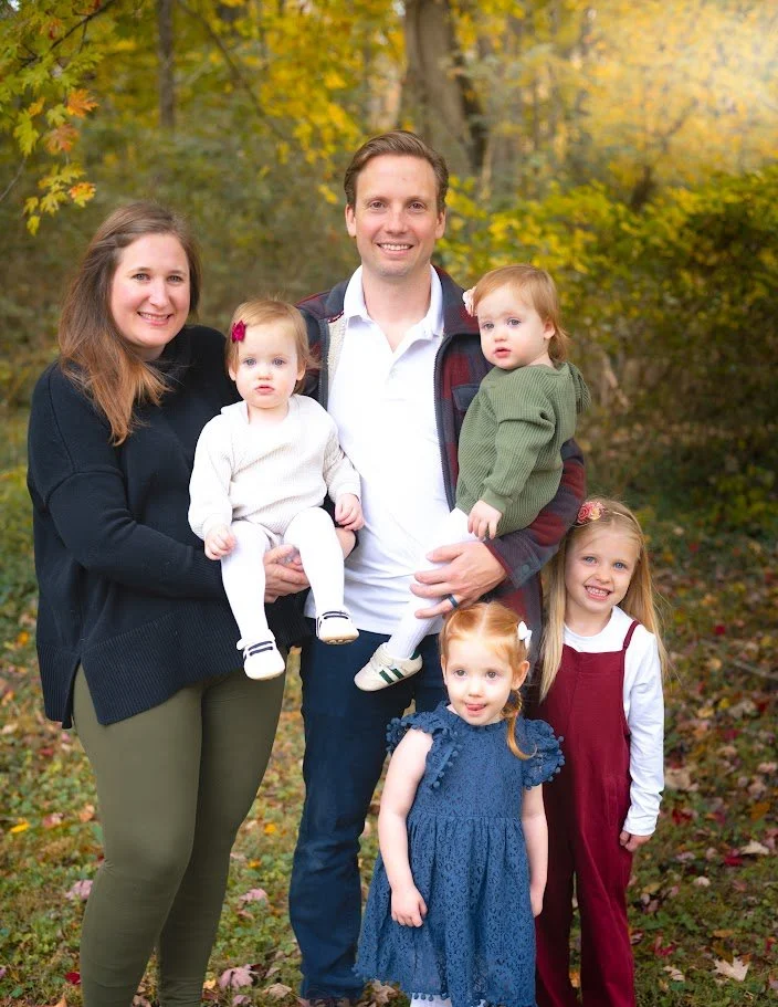 A family of six posing outdoors surrounded by fall foliage. The family consists of a woman, a man, and four young children, all smiling at the camera.