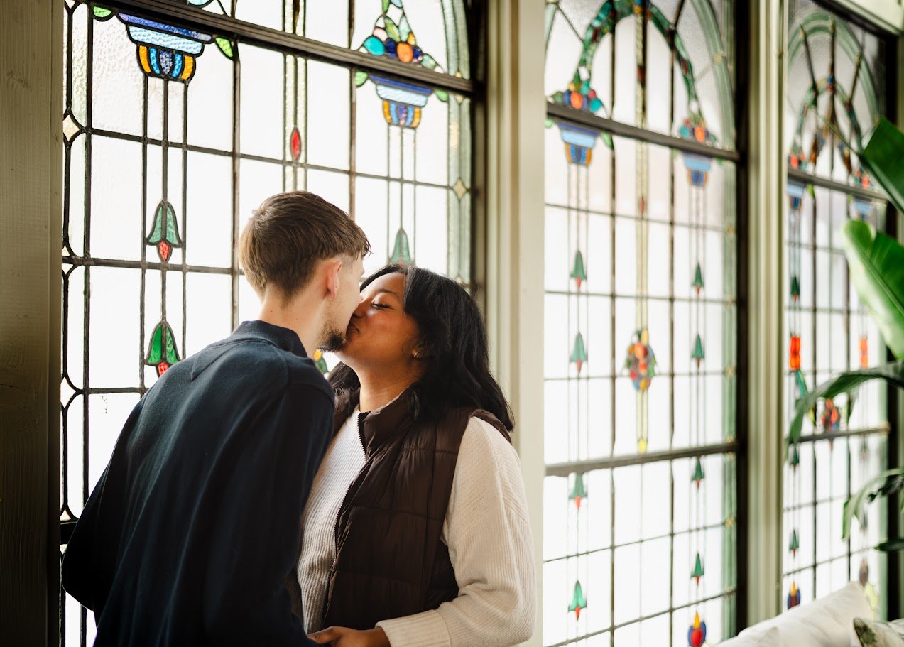 A couple kissing in front of stained glass windows with colorful floral patterns.