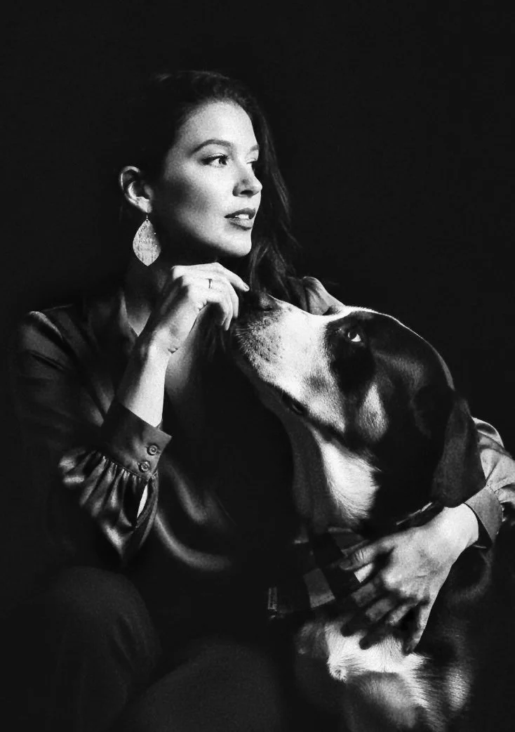 A woman with long dark hair wearing a satin blouse and large earrings, posing with a large dog with a black face and tan and white markings, against a dark background in a black and white photo.