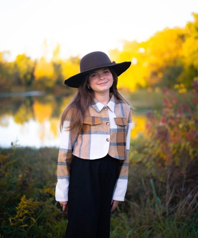 A young girl wearing a large black hat, a plaid jacket, and black pants standing outdoors near a lake during fall with trees showing yellow and red foliage.
