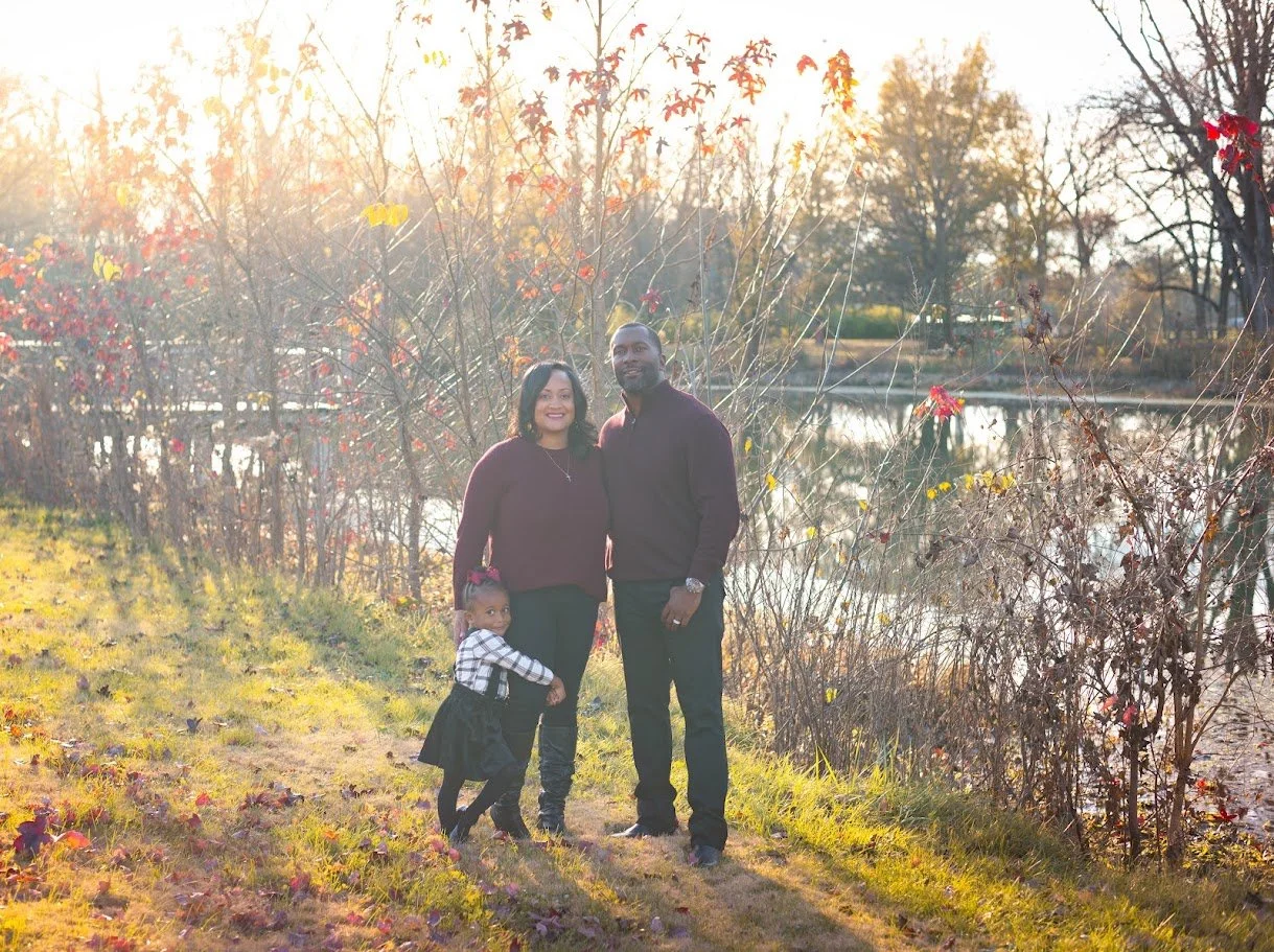 A family of three, including a woman, a man, and a young girl, stands outdoors in a park during autumn, near a body of water with trees and fall foliage in the background, illuminated by warm sunlight.