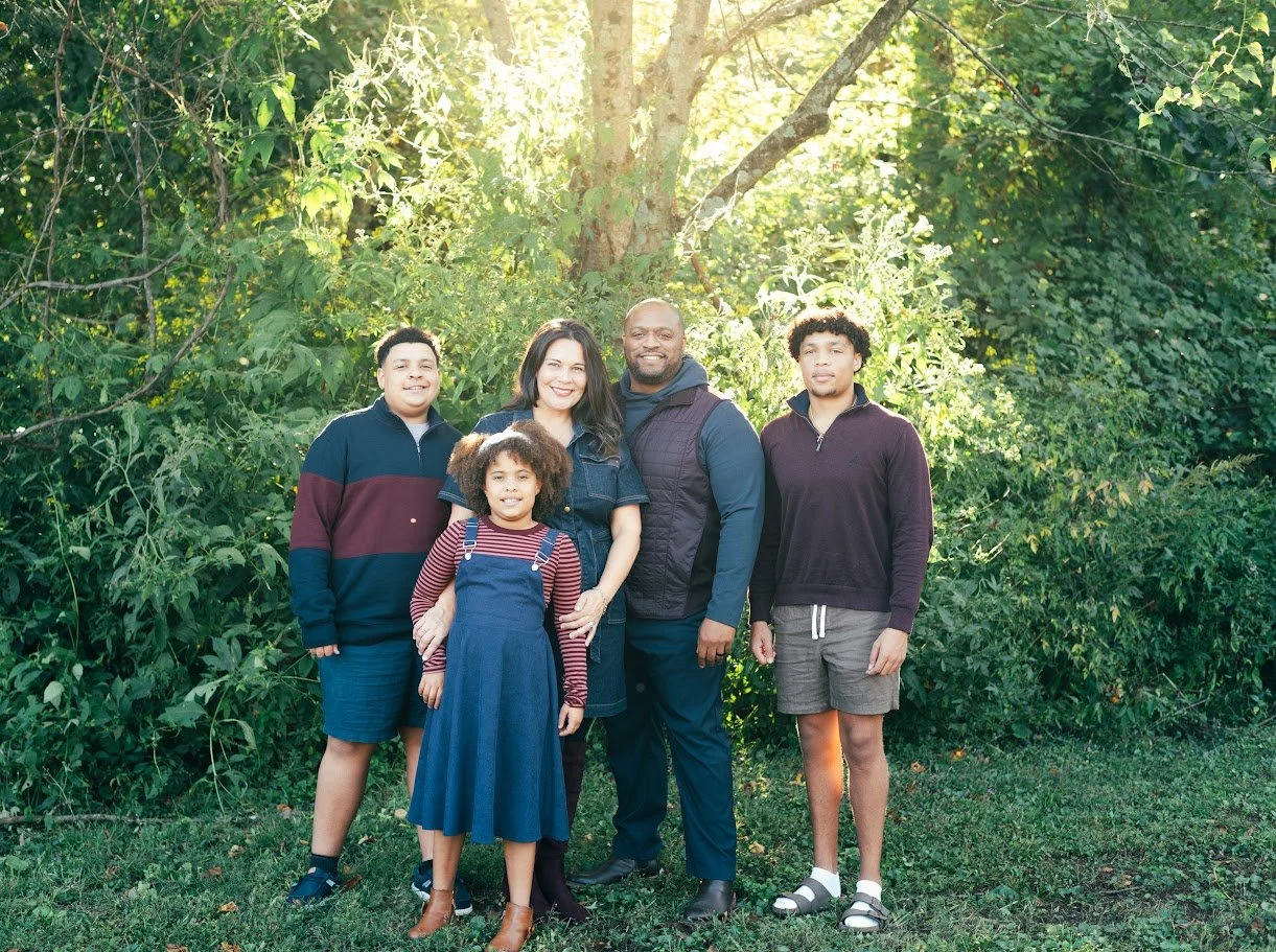 Family of five standing outdoors in front of lush green foliage, sunny weather.