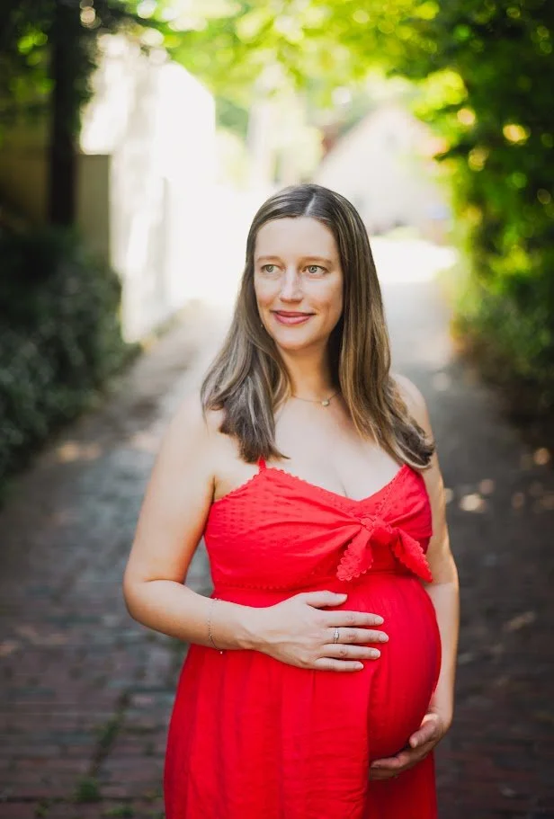 A pregnant woman in a red dress standing outdoors on a brick path surrounded by greenery, smiling and cradling her belly.