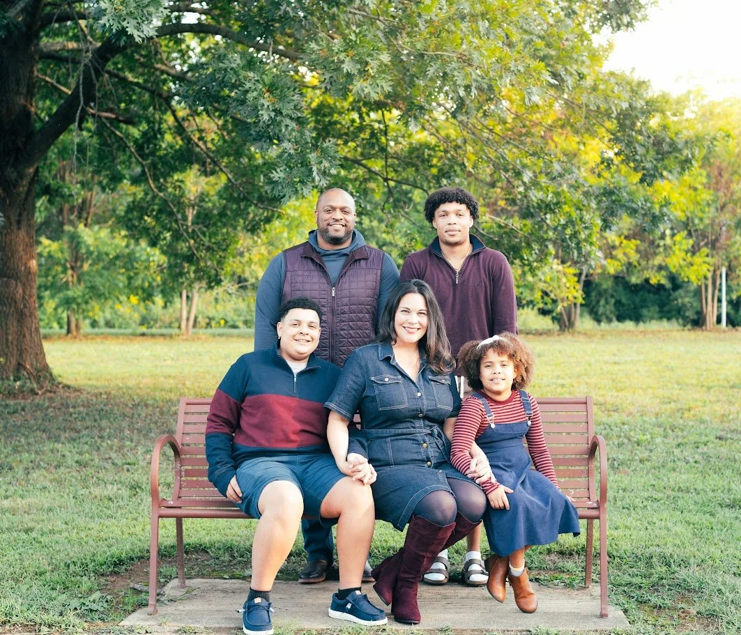 A family of six sitting on a park bench under a large tree, smiling at the camera. It includes a woman, two young boys, a young girl, and two men standing behind the bench.