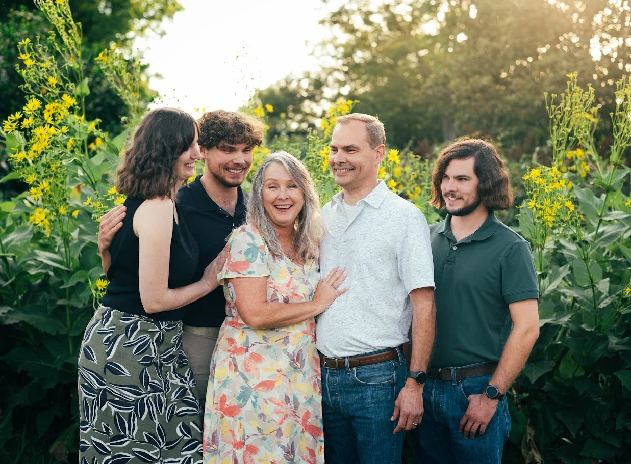 A group of five people smiling and standing close together outdoors among green plants and yellow flowers, with sunlight in the background.