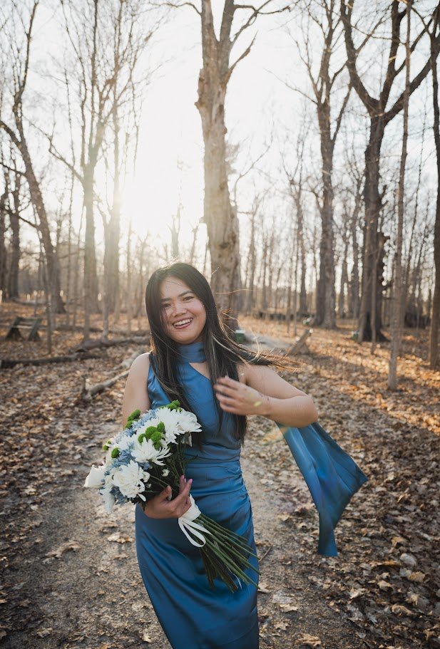 A woman in a blue dress holding a bouquet of white and green flowers, smiling and walking through a wooded area during late afternoon or early evening with bare trees and fallen leaves.