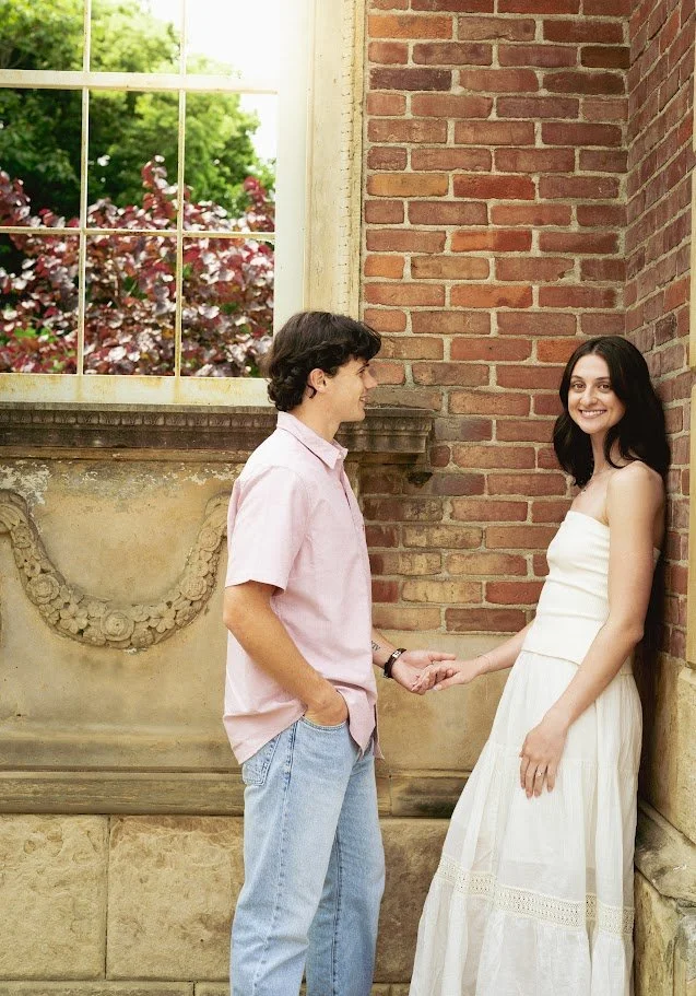 A young man and woman holding hands indoors near a brick wall, with a large window showing trees outside.