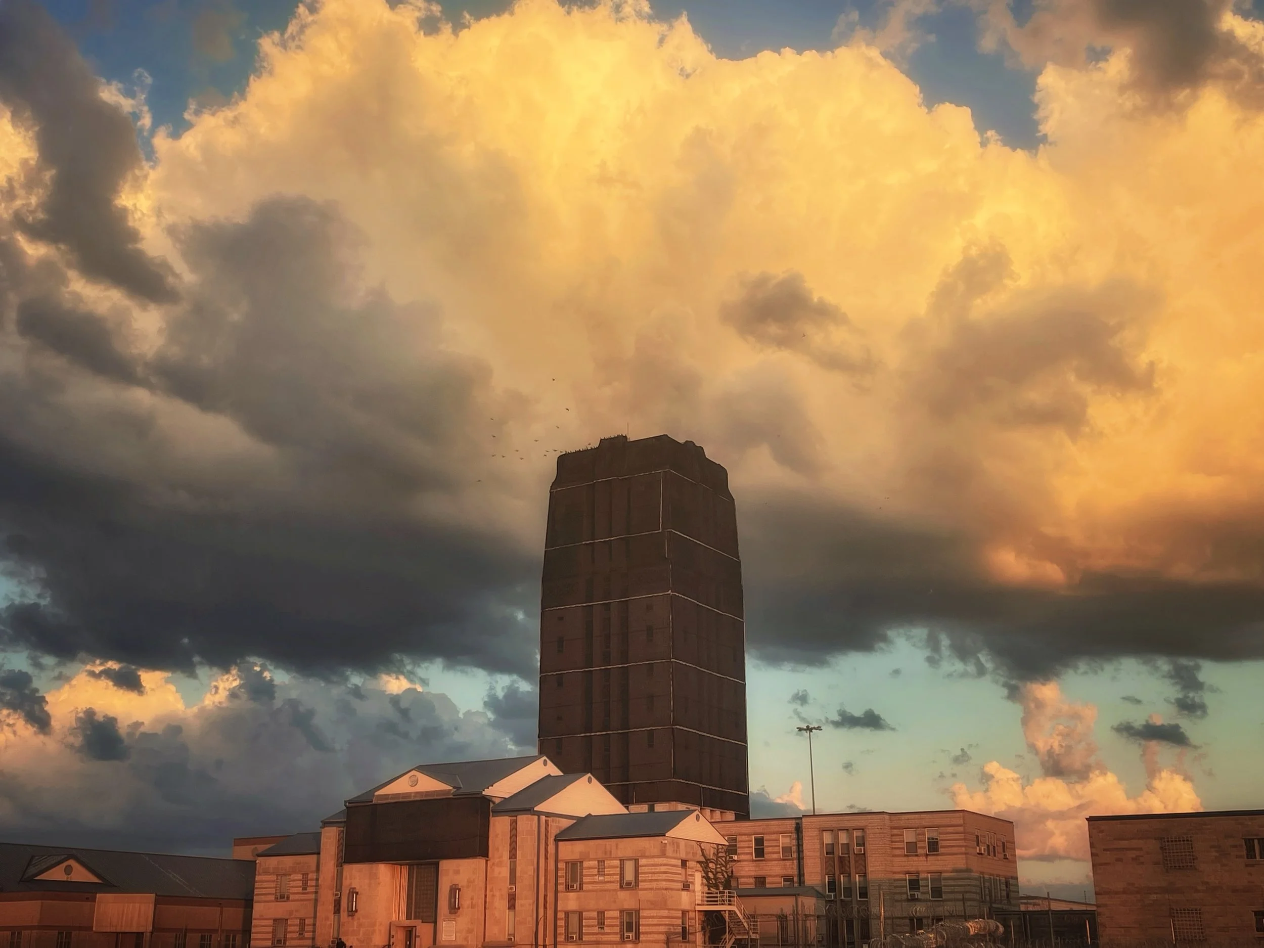 A tall brick building, likely a church or historic structure, standing against a sunset sky with clouds illuminated in shades of yellow, orange, and gray, surrounded by modern residential and commercial buildings.