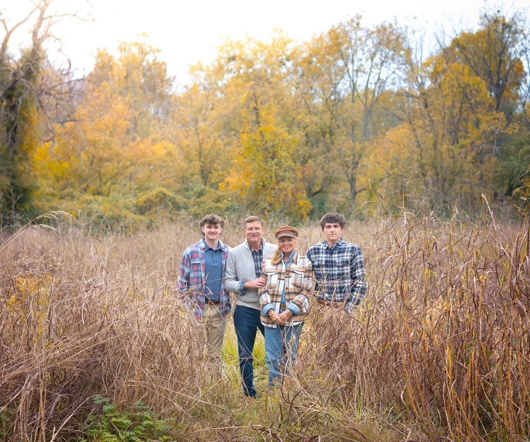 Family of four standing in a grassy field during autumn with colorful fall trees in the background.