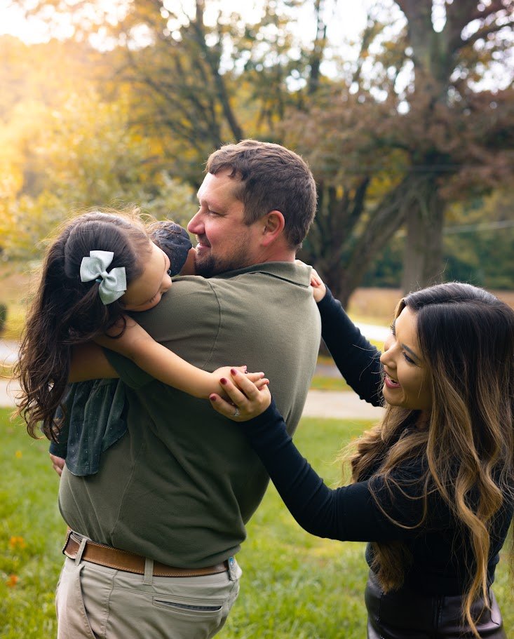 Family outdoors, father holding daughter, mother happily touching father's arm, autumn trees in background.